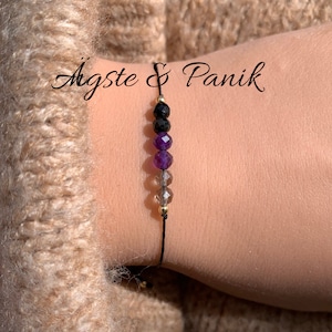 Könnte beinhalten: Ein schwarzes Armband aus Schnur mit schwarzen, violetten und braunen Perlen. Das Armband hat goldene Akzente und der Text "Agtste & Panik" ist im Hintergrund sichtbar.