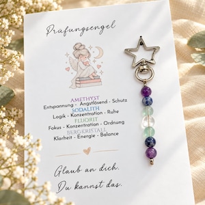 Könnte beinhalten: Ein sternförmiger Metallschlüsselanhänger mit einer Reihe aus violetten, blauen und klaren Perlen auf einer weißen Karte. Die Karte zeigt eine Engelsillustration, den Text Prüfungsengel, Kristallbeschreibungen sowie den Text Glaub an dich. Du kannst das.