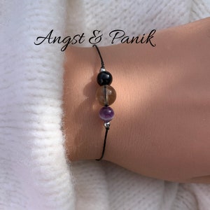 Könnte beinhalten: Schwarzes, braunes und violettes Perlenarmband auf einem schwarzen Band. Das Armband trägt den Text "Angst & Panik" darüber.