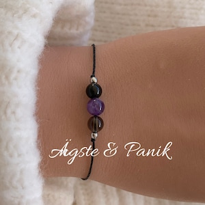 May include: A black string bracelet with three beads: black, amethyst, and smoky quartz. The bracelet has silver metal accents.