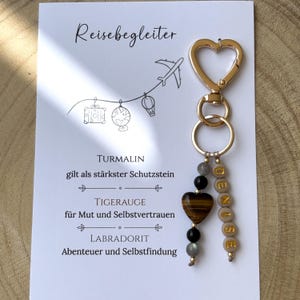 Könnte beinhalten: Ein goldfarbener herzförmiger Schlüsselanhänger mit einer herzförmigen Tigerauge-Perle, schwarzen und grauen Steinperlen sowie einer vertikalen Reihe beiger Buchstabenperlen, die DENISE bilden. Der Anhänger ist auf einer weißen Karte befestigt.
