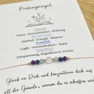 Può includere: Un braccialetto di perline con perline viola, blu, verdi e trasparenti, infilate su un sottile cordoncino beige. Il braccialetto è esposto accanto a una carta con testo in tedesco, inclusa la parola "Prüfungsengel".