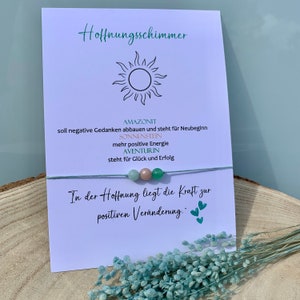 Peut inclure: Un bracelet composé de perles vertes, roses et vert clair avec une carte qui dit "Hoffnungsschimmer" et comprend le texte "In der Hoffnung liegt die Kraft zur positiven Veränderung." La carte liste également les pierres et leurs significations : Amazonite, Sunstone et Aventurine.