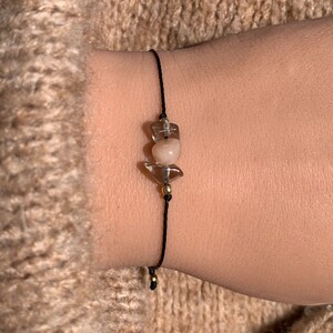 May include: A black string bracelet with three clear and pink crystal beads. The beads are tied together with gold wire.