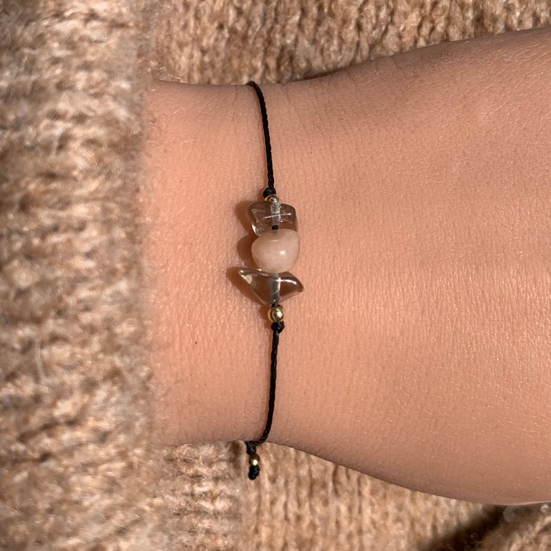 May include: A black string bracelet with three clear and pink crystal beads. The beads are tied together with gold wire.