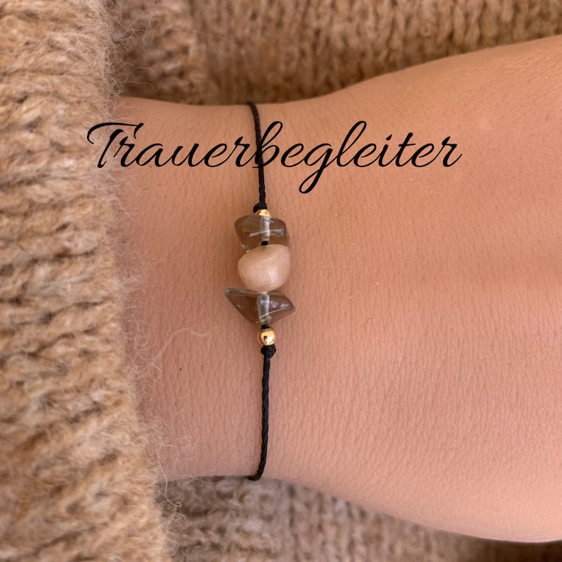 May include: A black string bracelet with three brown and beige gemstone beads. The bracelet has the word "Trauerbegleiter" written on it.