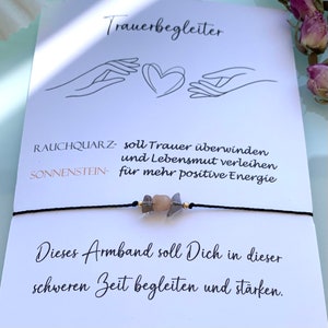 May include: A black string bracelet with two light brown and gray stones and gold beads. The bracelet is on a white card with the text "Trauerbegleiter", "RAUCHQUARZ- soll Trauer &uuml;berwinden und Lebensmut verleihen", "SONNENSTEIN- f&uuml;r mehr positive Energie", "Dieses Armband soll Dich in dieser schweren Zeit begleiten und st&auml;rken."