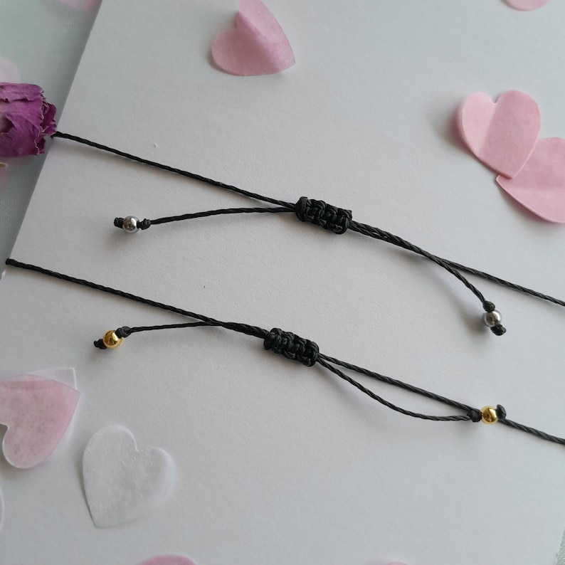 May include: Two black cord bracelets with silver and gold beads. The bracelets are tied with a knot at the end.