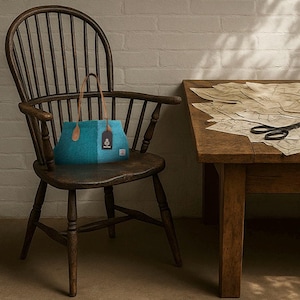 May include: A teal tote bag with brown leather handles and a tag, on a dark wooden chair. The bag has a two-tone design and a rectangular shape. The chair is near a wooden table with sewing patterns.