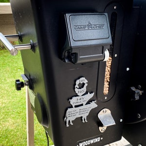 Puede incluir: Una parrilla de pellets negra Camp Chef Woodwind con un indicador de temperatura de metal plateado y una gu&iacute;a de temperatura de carne de metal plateado en forma de cerdo y vaca. La gu&iacute;a muestra las temperaturas internas recomendadas para diferentes tipos de carne.