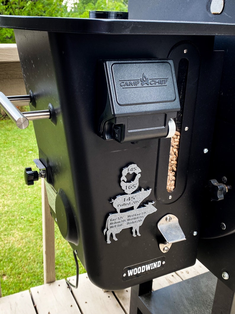 Puede incluir: Una parrilla de pellets negra Camp Chef Woodwind con un indicador de temperatura de metal plateado y una gu&iacute;a de temperatura de carne de metal plateado en forma de cerdo y vaca. La gu&iacute;a muestra las temperaturas internas recomendadas para diferentes tipos de carne.