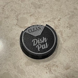 Puede incluir: Un "Dish Pal" redondo negro y gris con las palabras "Dish Pal" y "Clean" en &eacute;l.