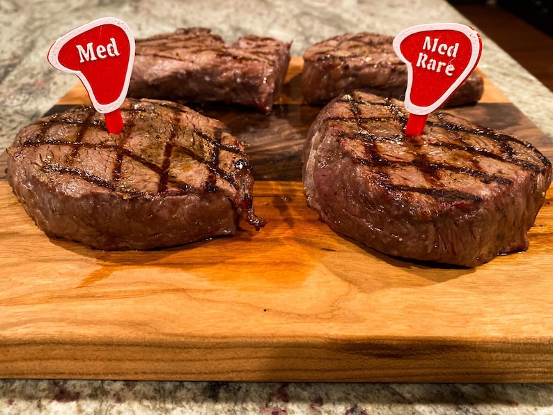 Puede incluir: Cuatro filetes a la parrilla en una tabla de cortar de madera. Dos filetes est&aacute;n etiquetados con "Med" y "Med Rare" con banderas rojas.