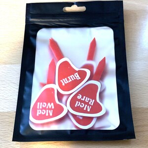 Puede incluir: Una bolsa de pl&aacute;stico transparente con cierre de cremallera negra contiene seis marcadores de carne en forma de coraz&oacute;n rojo. Los marcadores est&aacute;n etiquetados como "A1cd Well", "A1cd Rare" y "Burnt".