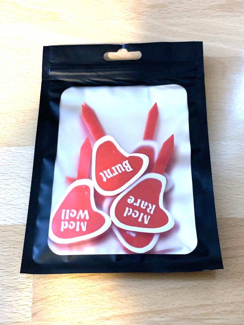Puede incluir: Una bolsa de pl&aacute;stico transparente con cierre de cremallera negra contiene seis marcadores de carne en forma de coraz&oacute;n rojo. Los marcadores est&aacute;n etiquetados como "A1cd Well", "A1cd Rare" y "Burnt".
