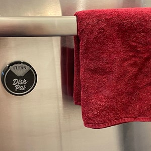 Puede incluir: Un im&aacute;n redondo negro para el seguimiento del secado de los platos con las palabras "Clean" y "Dish Pal" en &eacute;l. El im&aacute;n est&aacute; fijado a una puerta de refrigerador de acero inoxidable con una toalla roja colgada en una barra encima.