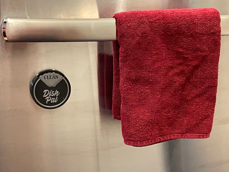 Puede incluir: Un im&aacute;n redondo negro para el seguimiento del secado de los platos con las palabras "Clean" y "Dish Pal" en &eacute;l. El im&aacute;n est&aacute; fijado a una puerta de refrigerador de acero inoxidable con una toalla roja colgada en una barra encima.