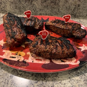 Puede incluir: Cuatro filetes a la parrilla en un plato rojo con un patr&oacute;n de estrellas blancas y negras. Los filetes est&aacute;n etiquetados con banderas rojas que indican su nivel de cocci&oacute;n: poco hecho, poco hecho y medio.