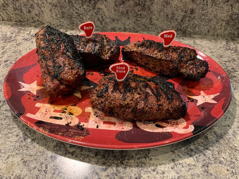 Puede incluir: Cuatro filetes a la parrilla en un plato rojo con un patr&oacute;n de estrellas blancas y negras. Los filetes est&aacute;n etiquetados con banderas rojas que indican su nivel de cocci&oacute;n: poco hecho, poco hecho y medio.