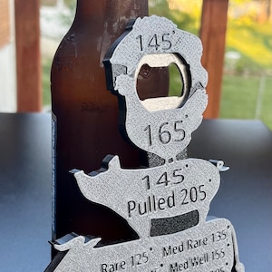 May include: A silver metal bottle opener shaped like a cow with different internal temperatures for cooking meat. The temperatures are listed for rare, medium rare, medium, medium well, well, and pulled pork. The bottle opener is next to a brown glass beer bottle and a silver bottle cap.