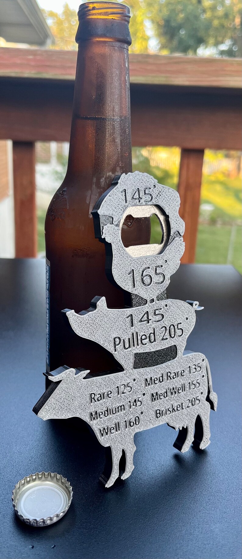 May include: A silver metal bottle opener shaped like a cow with different internal temperatures for cooking meat. The temperatures are listed for rare, medium rare, medium, medium well, well, and pulled pork. The bottle opener is next to a brown glass beer bottle and a silver bottle cap.
