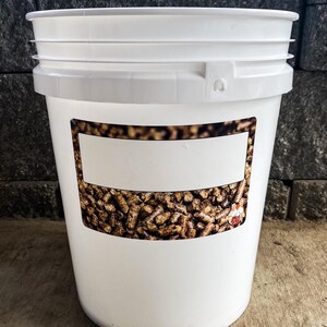 Pu&ograve; includere: Un secchio di plastica bianco con coperchio. Il secchio ha un'etichetta rettangolare con uno sfondo strutturato marrone e beige. Il secchio &egrave; appoggiato su una superficie di legno.