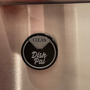 Puede incluir: Un im&aacute;n redondo para lavavajillas negro y plateado con el texto "Dish Pal" y "CLEAN" en &eacute;l.