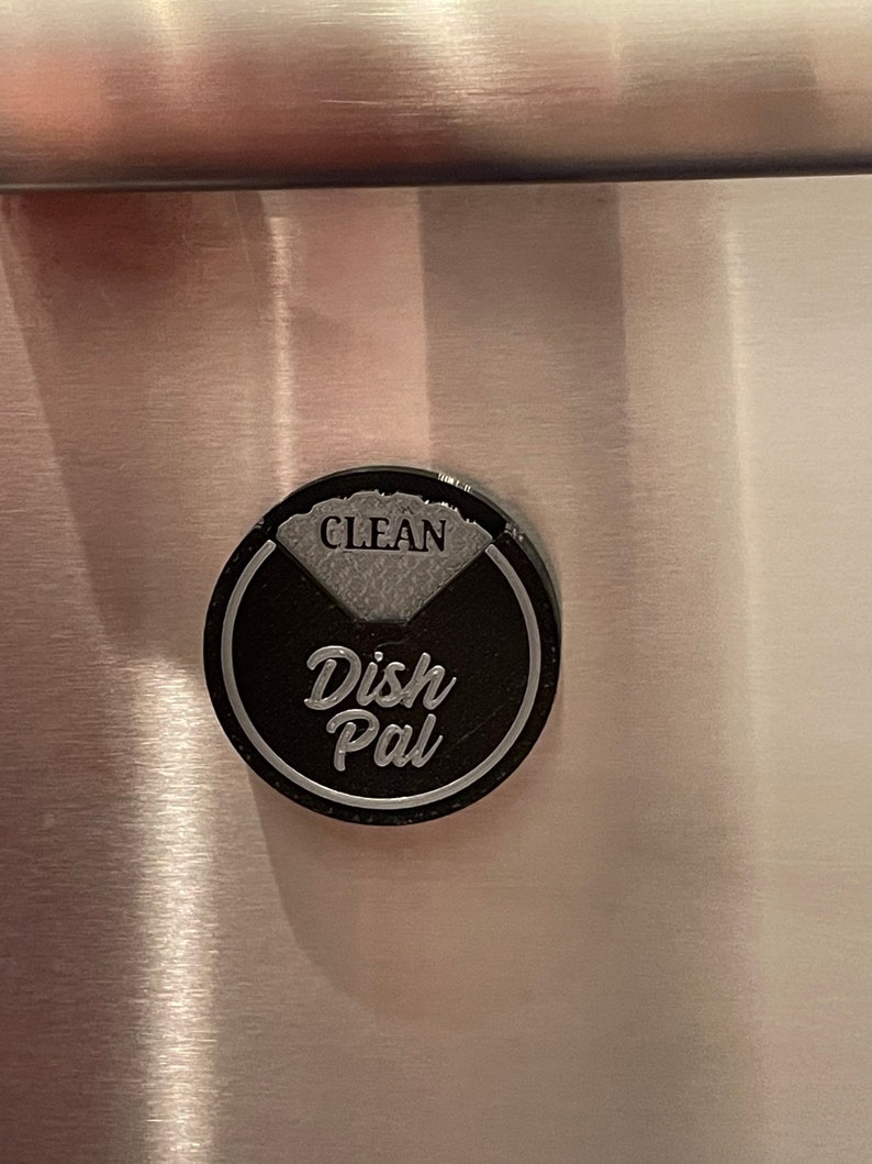 Puede incluir: Un im&aacute;n redondo para lavavajillas negro y plateado con el texto "Dish Pal" y "CLEAN" en &eacute;l.