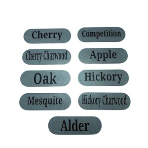 May include: Nine silver oval labels with black text listing different wood types: Cherry, Cherry Charwood, Oak, Mesquite, Alder, Competition, Apple, Hickory, Hickory Charwood.