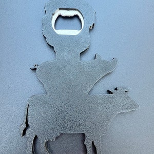 May include: Black metal bottle opener shaped like a cow with a pig on its back and a person on top of the pig. The person has a bottle opener in their hand.