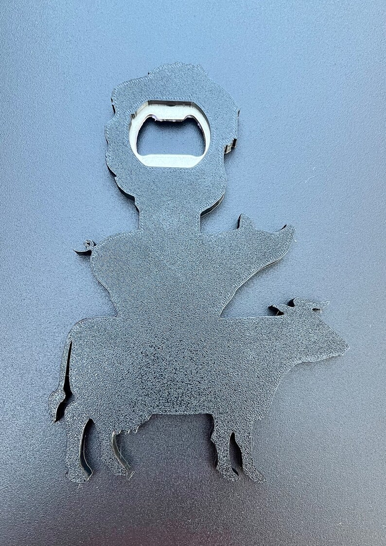 May include: Black metal bottle opener shaped like a cow with a pig on its back and a person on top of the pig. The person has a bottle opener in their hand.