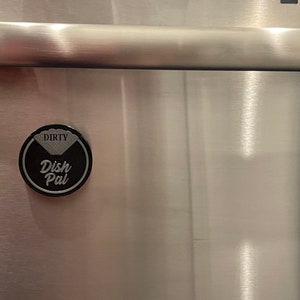 Puede incluir: Un im&aacute;n redondo negro con las palabras "Dish Pal" y "Dirty" en &eacute;l. El im&aacute;n est&aacute; pegado a un refrigerador de acero inoxidable.