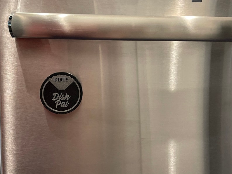 Puede incluir: Un im&aacute;n redondo negro con las palabras "Dish Pal" y "Dirty" en &eacute;l. El im&aacute;n est&aacute; pegado a un refrigerador de acero inoxidable.