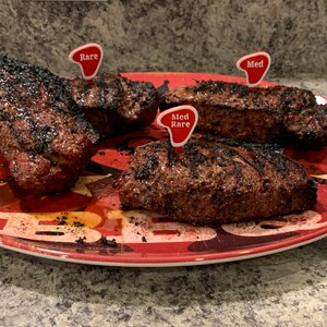 Puede incluir: Tres filetes a la parrilla en un plato rojo y negro con un dise&ntilde;o de estrellas. Los filetes est&aacute;n etiquetados con marcadores de pl&aacute;stico rojo que indican "Rare", "Med Rare" y "Med".