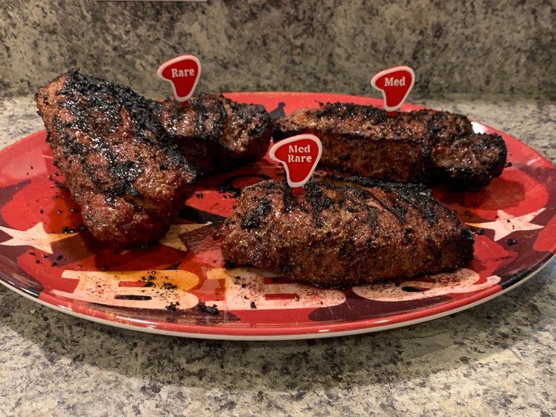 Puede incluir: Tres filetes a la parrilla en un plato rojo y negro con un dise&ntilde;o de estrellas. Los filetes est&aacute;n etiquetados con marcadores de pl&aacute;stico rojo que indican "Rare", "Med Rare" y "Med".