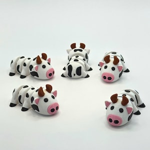 May include: A collection of six small, white cow figurines with black spots, pink noses, and brown horns. The figurines are arranged on a white surface, with some lying down and others upright. The overall theme is farm animals.