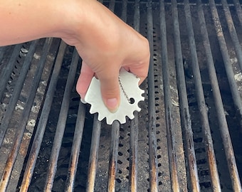 May include: A close-up of a hand using a grill cleaning tool to scrub a dirty grill grate. The tool is a star-shaped metal scrubber with a chain attached.