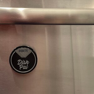 Puede incluir: Un im&aacute;n redondo negro y plateado con el texto "Dirty Dish Pal" en &eacute;l. El im&aacute;n est&aacute; fijado a una superficie de acero inoxidable.