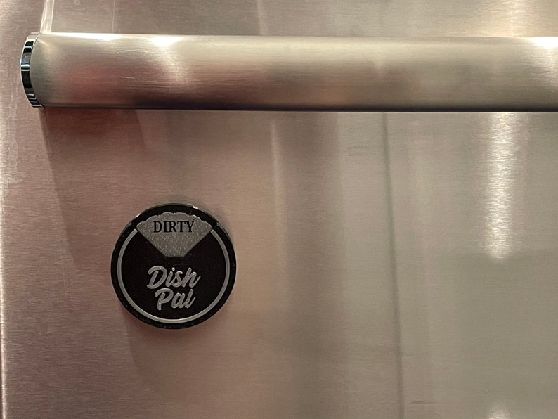 Puede incluir: Un im&aacute;n redondo negro y plateado con el texto "Dirty Dish Pal" en &eacute;l. El im&aacute;n est&aacute; fijado a una superficie de acero inoxidable.