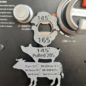 May include: A silver metal grill thermometer with a pig and cow design. The thermometer has a built-in bottle opener and displays different temperatures for various meats, including rare, medium rare, medium, medium well, well, and pulled pork. The thermometer also includes the text "IGNITOR ALLUMEUR" and "LOW bas bajo".