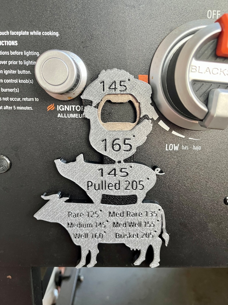 May include: A silver metal grill thermometer with a pig and cow design. The thermometer has a built-in bottle opener and displays different temperatures for various meats, including rare, medium rare, medium, medium well, well, and pulled pork. The thermometer also includes the text "IGNITOR ALLUMEUR" and "LOW bas bajo".