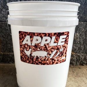 Pu&ograve; includere: Un secchio di plastica bianco con coperchio e manico in metallo. Il secchio ha un'etichetta con la parola "APPLE" a grandi lettere bianche. L'etichetta presenta anche illustrazioni di un pollo, un maiale e verdure.