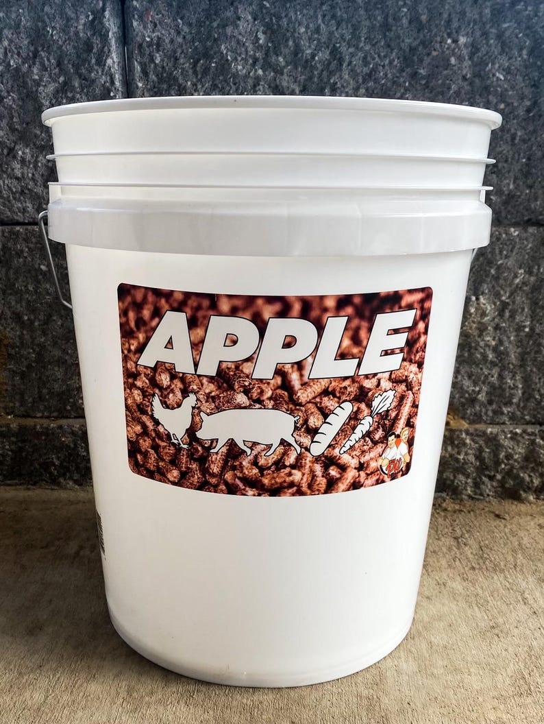 Pu&ograve; includere: Un secchio di plastica bianco con coperchio e manico in metallo. Il secchio ha un'etichetta con la parola "APPLE" a grandi lettere bianche. L'etichetta presenta anche illustrazioni di un pollo, un maiale e verdure.