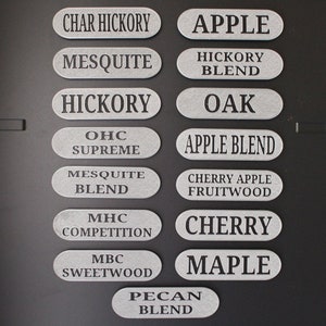 May include: A collection of 12 oval-shaped wood chips for smoking, each with a different wood type or blend printed on them. The wood types include: Char Hickory, Apple, Mesquite, Hickory Blend, Hickory, Oak, OHC Supreme, Apple Blend, Mesquite Blend, Cherry Apple Fruitwood, MHC Competition, MBC Sweetwood, Cherry, Maple, and Pecan Blend.