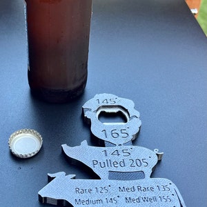 May include: A metal cow-shaped bottle opener with a built-in thermometer. The thermometer has markings for different temperatures, including rare, medium rare, medium, medium well, well, and pulled. The bottle opener is on a black surface with a bottle of beer and a bottle cap.