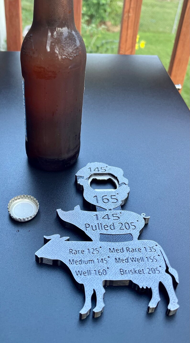 May include: A metal cow-shaped bottle opener with a built-in thermometer. The thermometer has markings for different temperatures, including rare, medium rare, medium, medium well, well, and pulled. The bottle opener is on a black surface with a bottle of beer and a bottle cap.