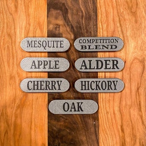 May include: Seven silver-colored, oval-shaped metal tags with engraved text, including "MESQUITE," "APPLE," "CHERRY," "COMPETITION BLEND," "ALDER," "HICKORY," and "OAK." The tags are arranged on a two-toned wooden surface.