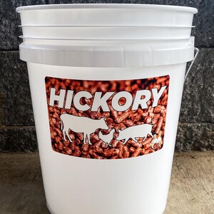 Pu&ograve; includere: Secchio bianco con coperchio e manico in metallo. Il secchio ha un'etichetta con la parola "HICKORY" in grandi lettere bianche. L'etichetta presenta anche sagome di una mucca, un pollo, un maiale e una piccola figura, su uno sfondo di pellet di legno marrone.