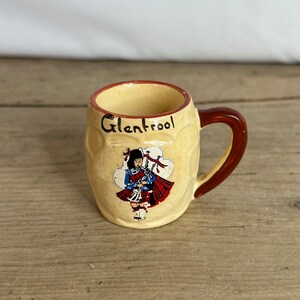May include: A small, cream-coloured ceramic mug with a brown handle and rim. The mug features a Scottish themed illustration of a person in traditional attire, with the word "Glentrool" printed on the front.