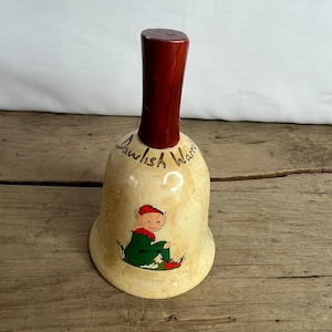 May include: Cream-colored ceramic bell with a dark red wooden handle. The bell features a seated elf illustration in green, red, and flesh tones. The words "Dawlish Warren" are written in cursive on the bell.